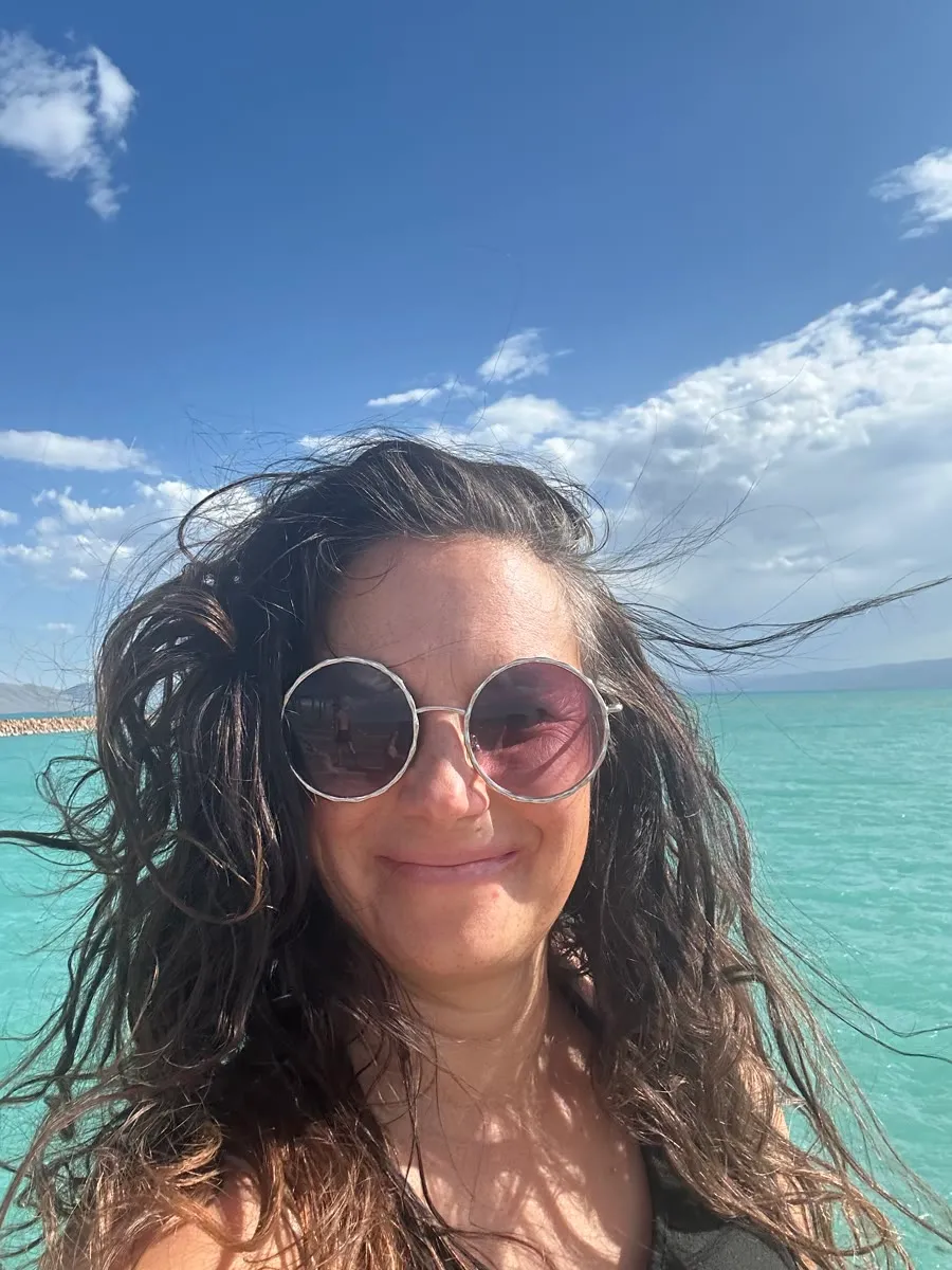 Vanessa today in round sunglasses, wind in her hair and turquoise water behind her.
