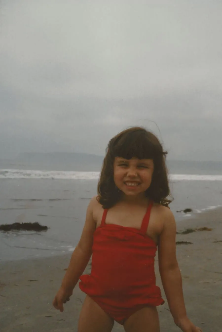 Vanessa as a young girl in a red swimsuit on an overcast beach.