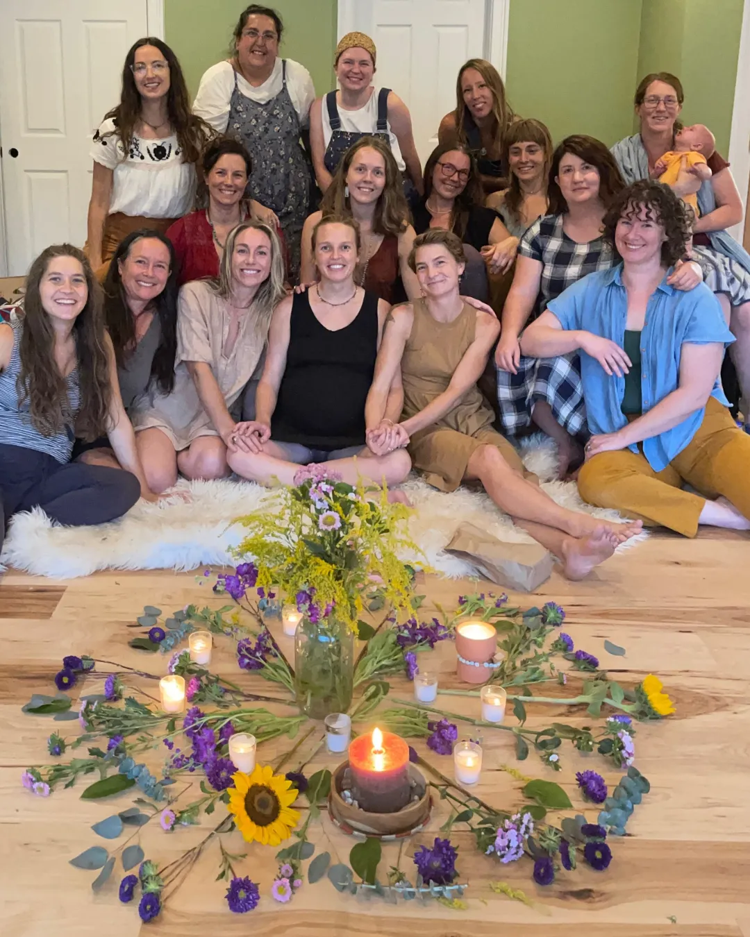 A circle of women seated together on the floor around a flower-and-candle altar.