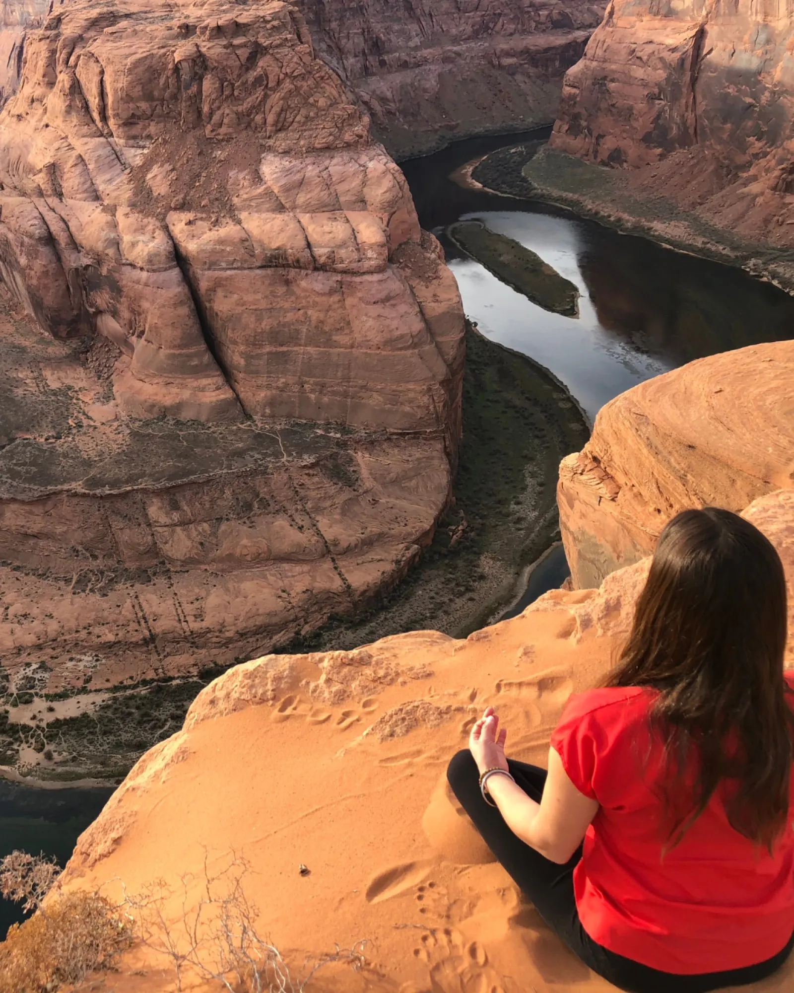 A woman in a red shirt seated in meditation, her back to the camera, overlooking the Horseshoe Bend canyon.