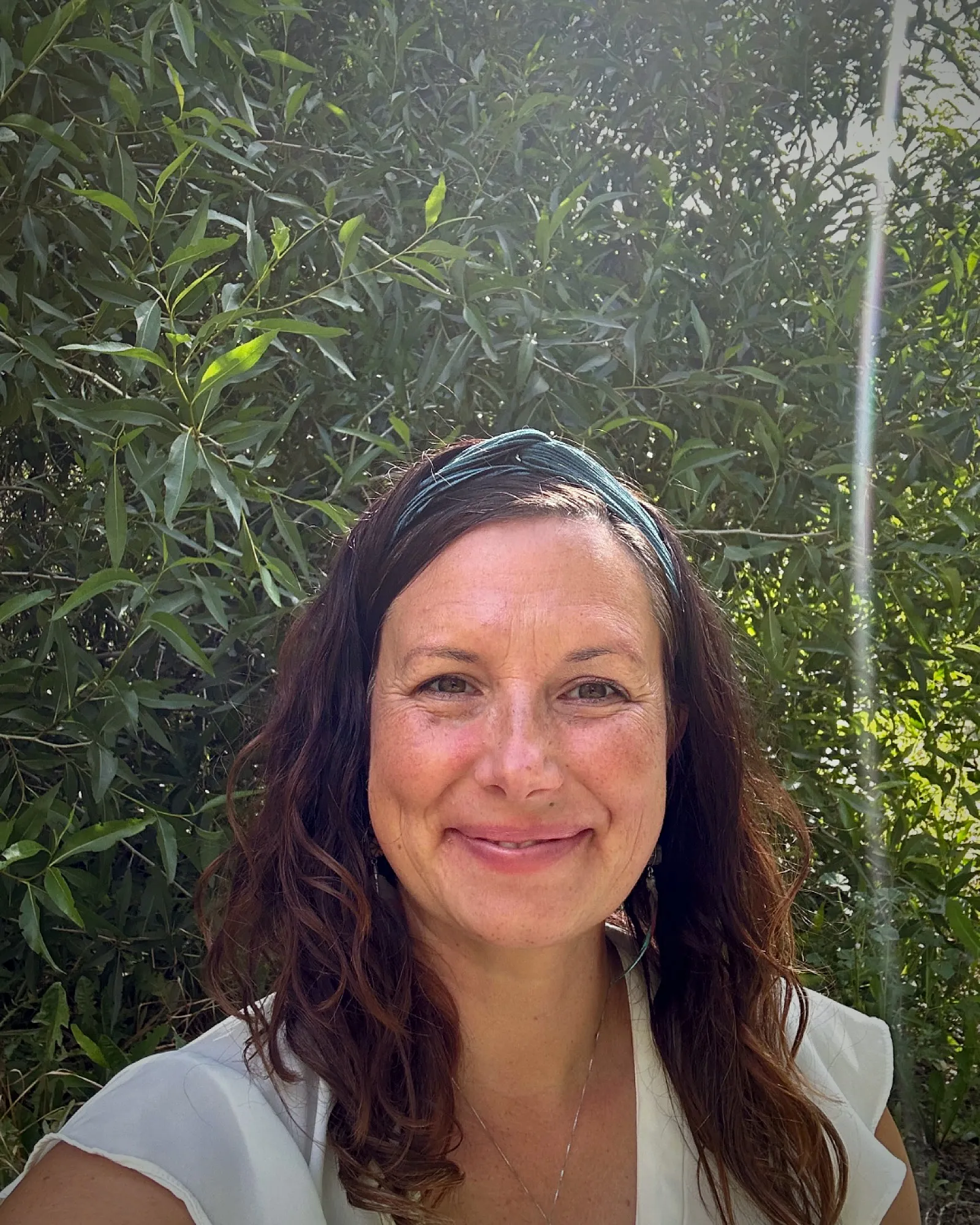 Vanessa, smiling warmly outdoors among green leaves.