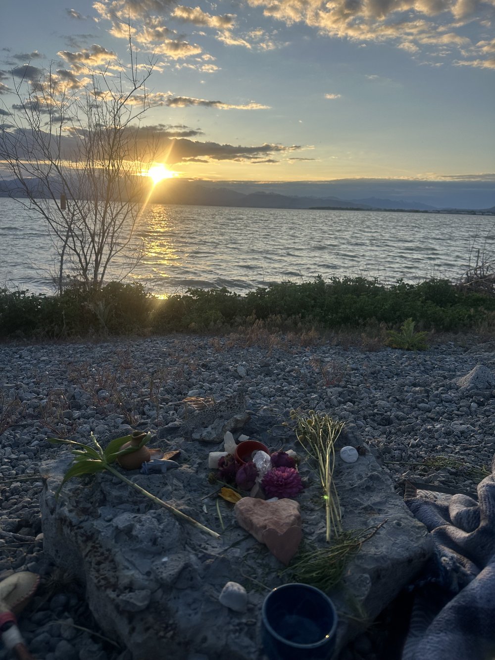 Our earth-vigil altar on the rocky bank at Utah Lake: garden clippings, dried flowers, crystals, a red bowl, a white candle, a heart-shaped stone, a moon-faced stone, and a blue handmade cup, with the sun setting over the water behind.