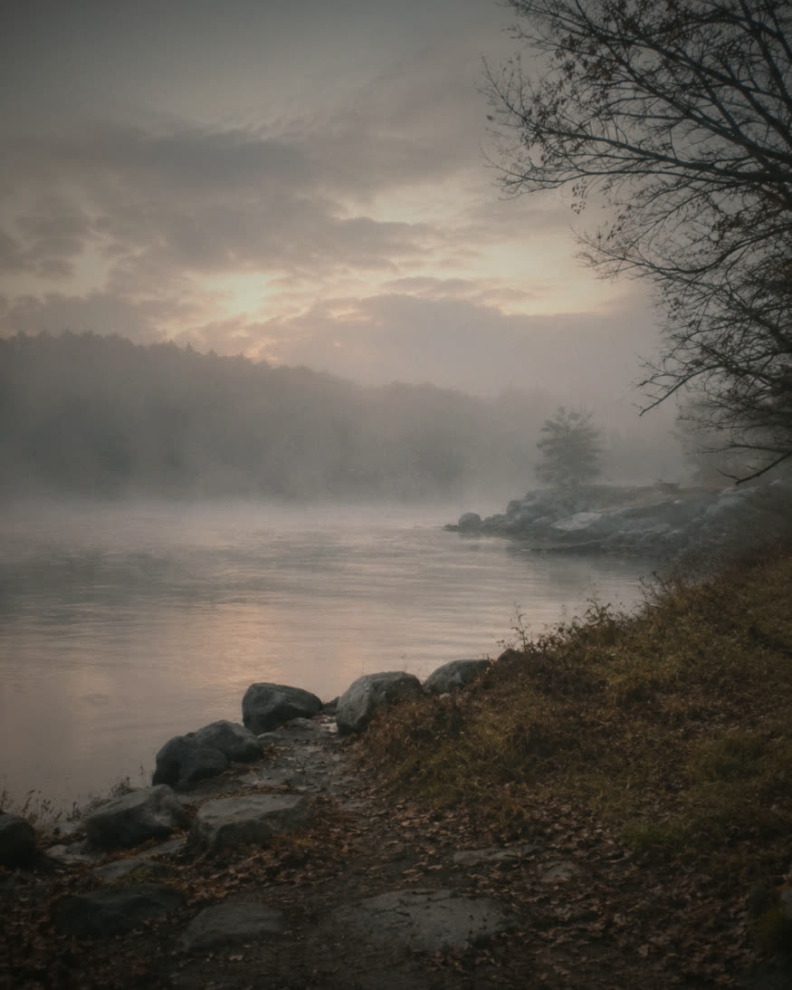 A still river at dawn, mist rising off the water above a rocky, leaf-strewn shore.